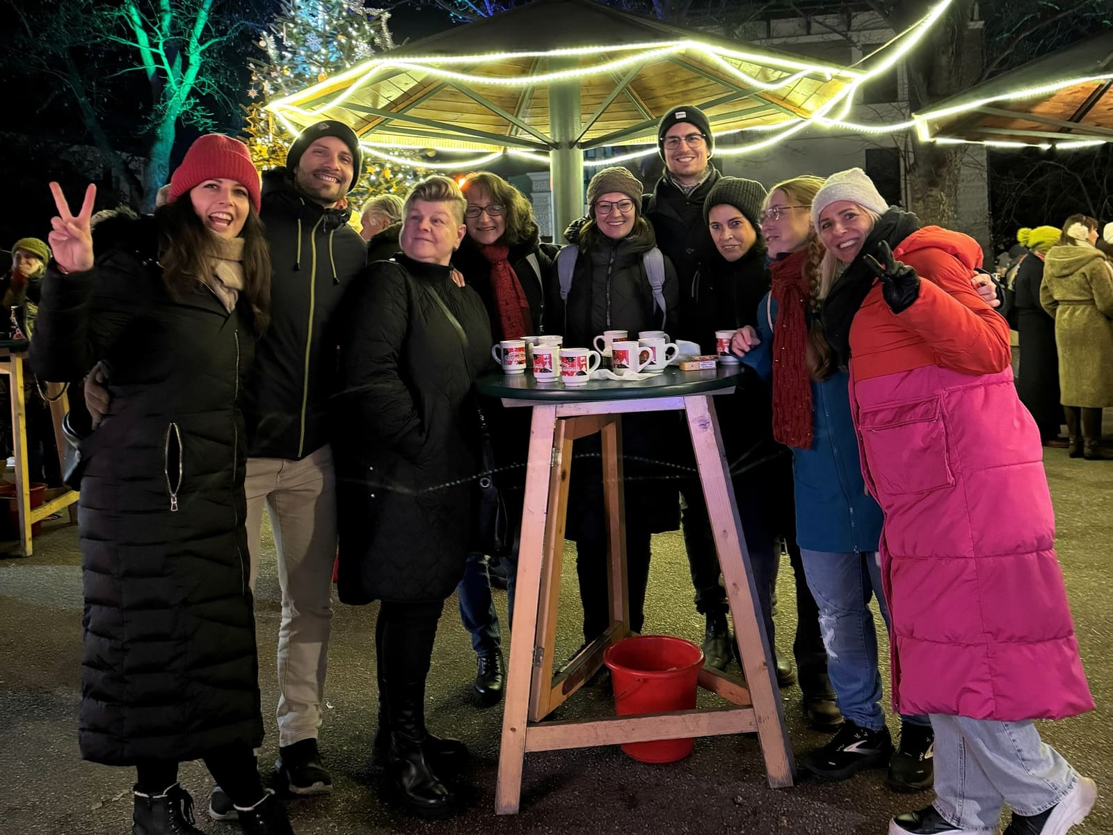 Neun Freunde stehen abends um einen Stehtisch mit Punschbechern auf einem Wiener Christkindlmarkt, dahinter beleuchtete Stände und Bäume.