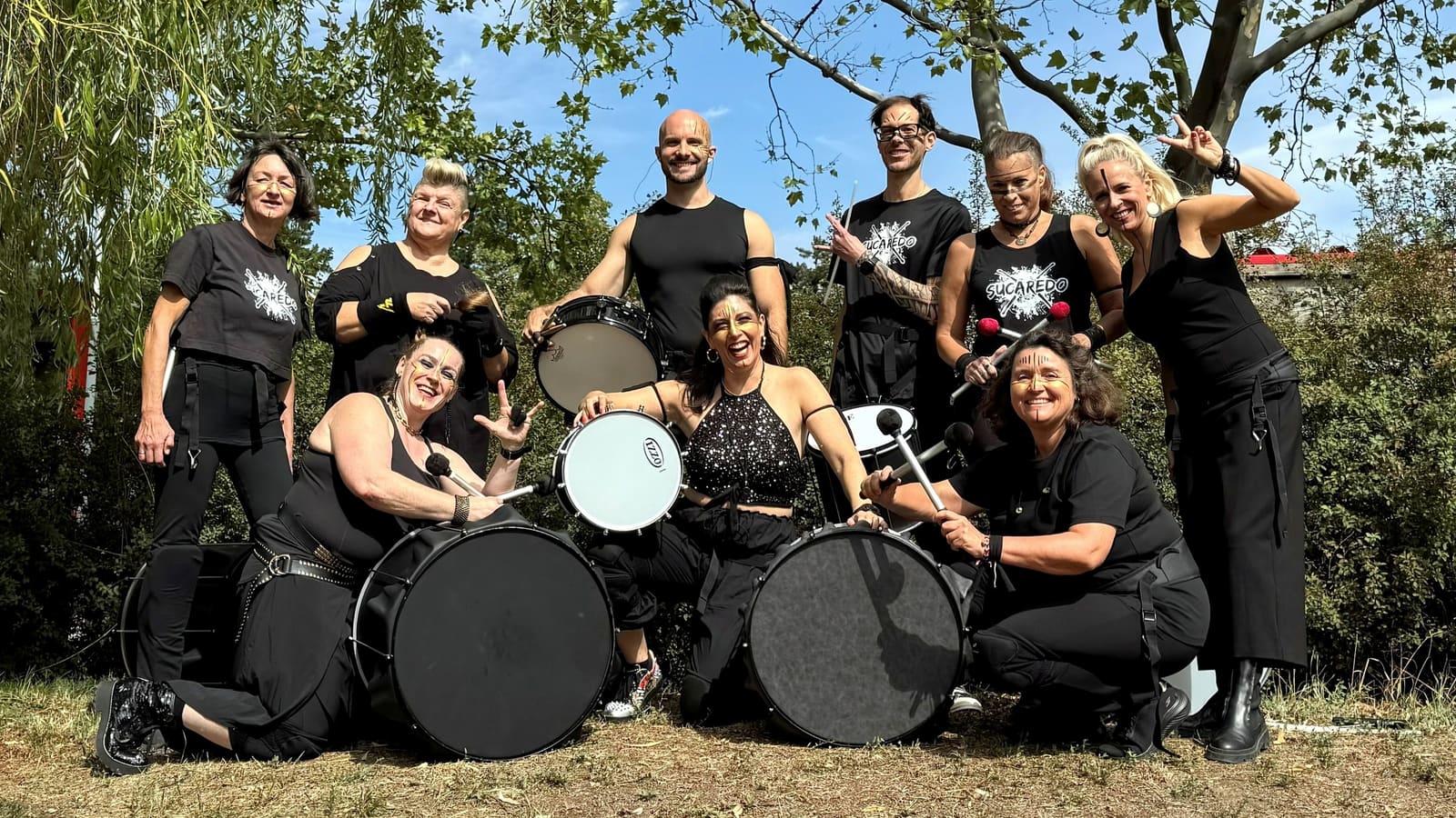 Neun Bandmitglieder von SUCAREDO in Schwarz posieren lachend mit ihren Trommeln und Sticks auf einer Wiese unter Bäumen.