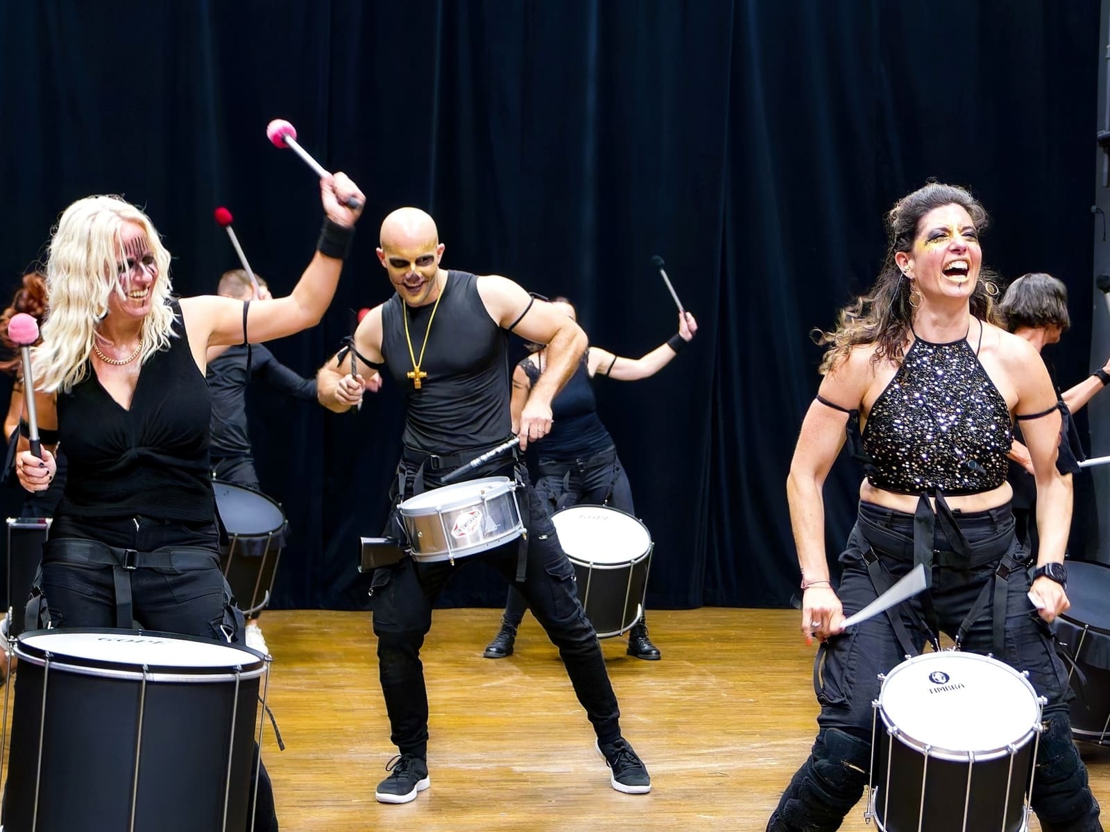 Fünf Bandmitglieder in Schwarz trommeln mit erhobenen Sticks auf einer Bühne vor dunklem Vorhang.