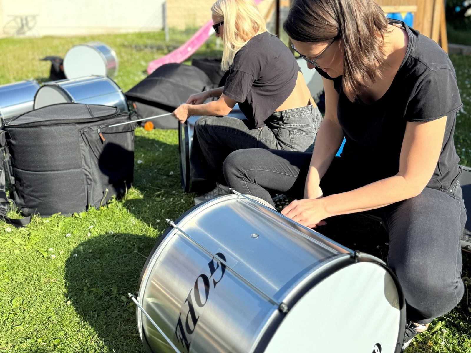 Zwei Frauen packen in einem sonnigen Garten neue, silbern glänzende Trommeln aus ihren Taschen aus.