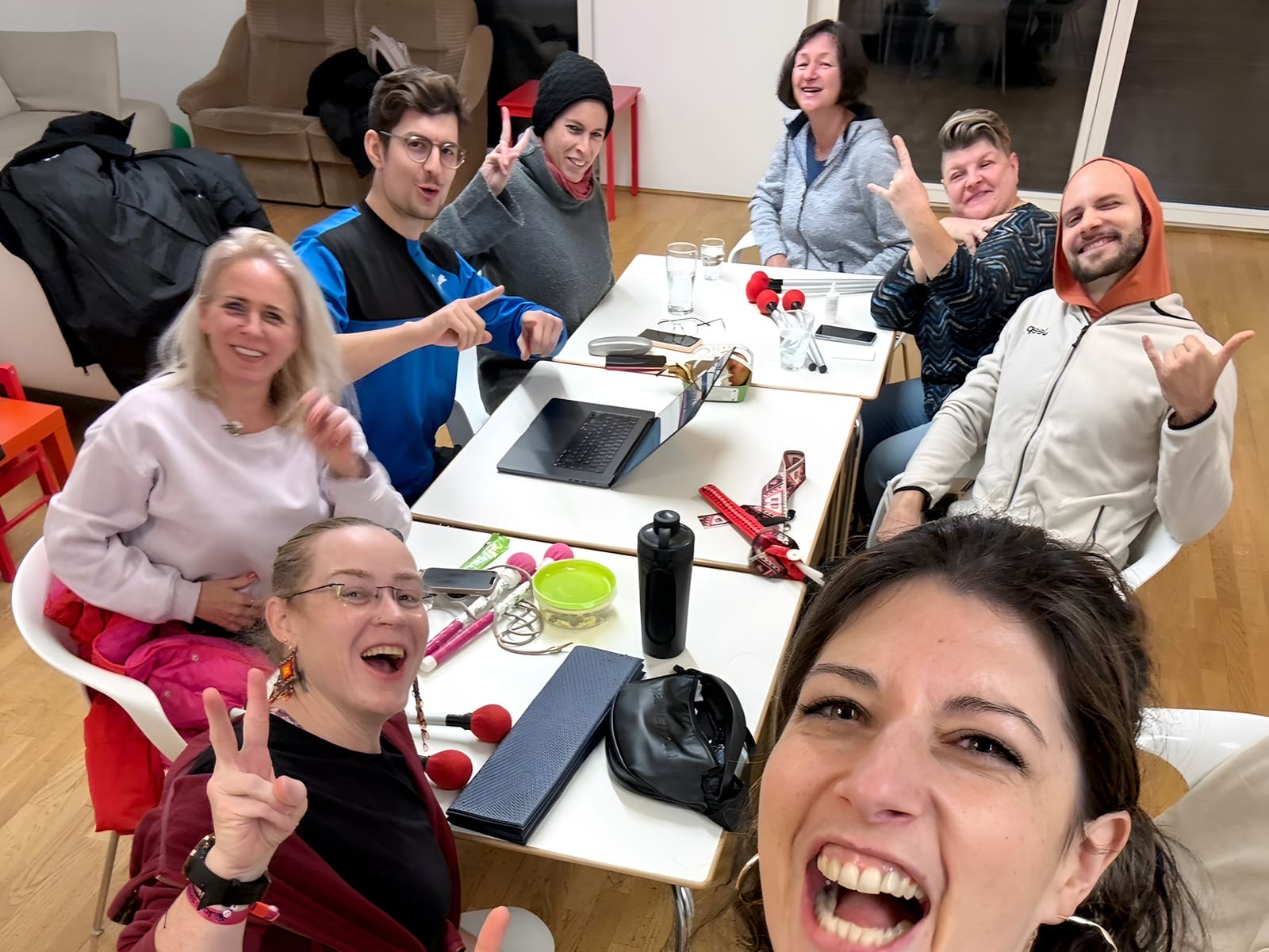 Selfie von acht lachenden Leuten an einem Tisch in einem Gemeinschaftsraum, auf dem Tisch Laptop, Sticks und Trinkflaschen.